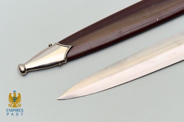 Close-up of a polished dagger with a metallic hilt and dark wooden grip on a pale surface; Empires Past logo bottom left.