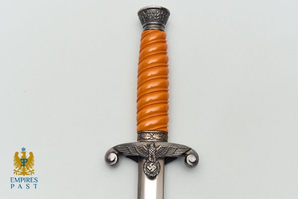 Close-up of a decorative dagger hilt with an orange spiral grip and ornate metal guard, against a pale background.