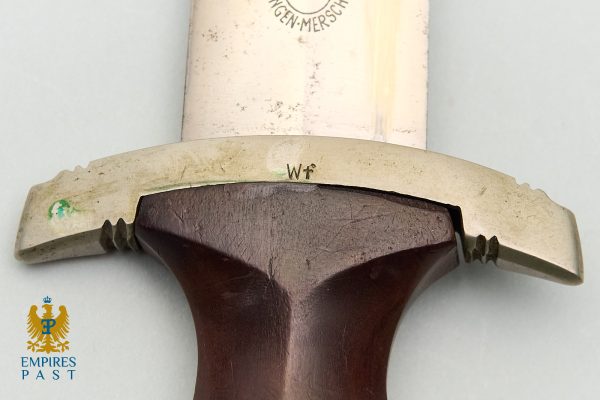 Close-up of a sword hilt: stamped 'Wf' on the crossguard, a brown wooden grip, and a worn blade edge.