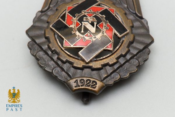 Metal badge with red and black enamel, gear wheel motif, central 'N' and 1922 on a ribbon, surrounded by laurel leaves
