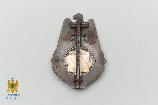 Antique metal badge with a circular emblem in the center, mounted on a shield-shaped backing; a rusted pin crosses the badge vertically.