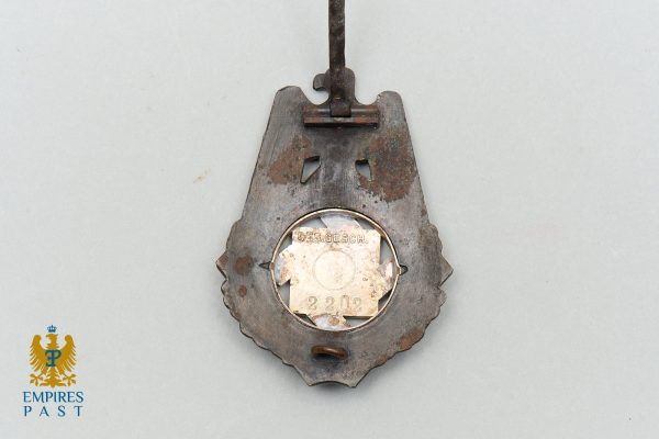 Rusty shield-shaped metal badge with a circular plate reading GES.GESCH and the number 2202, suspended from a cord. A faded emblems logo appears bottom-left.