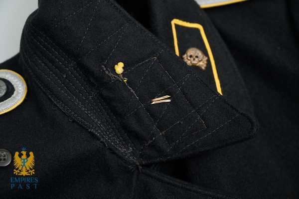 Close-up of a black wool military jacket collar with yellow piping, a tiny yellow boutonniere, and two small metal pins.