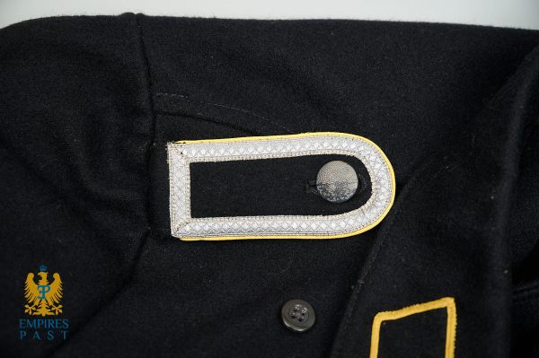 Close-up of a black military uniform with a silver-bordered, yellow-edged epaulet and a gray button.