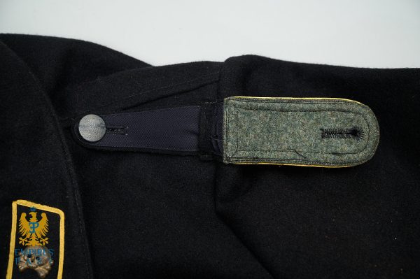 Close-up of a dark uniform jacket shoulder with a gray fabric strap and a metal snap, plus a padded green loop; a gold-edged crest patch with an eagle is visible on the left.