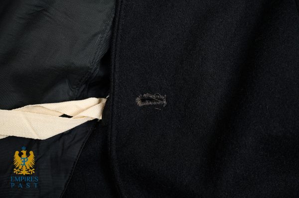 Close-up of a black wool coat showing a small frayed buttonhole and a beige fabric strap tucked into the seam, with the Empire’s Past logo in the lower-left corner.