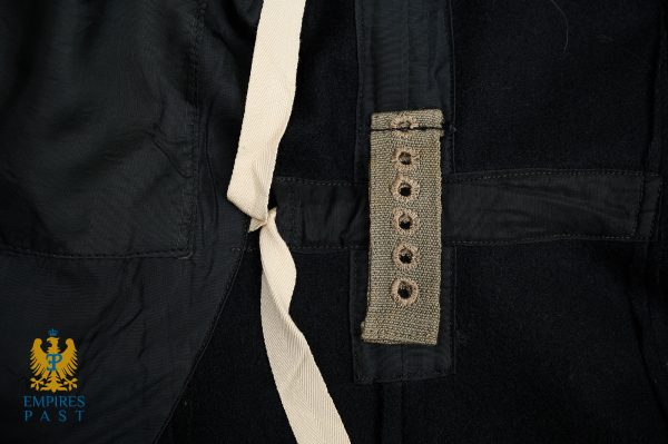Close-up of a dark fabric uniform with a beige strap and a hooked belt loop, plus a small blue-and-gold insignia in the bottom-left corner.