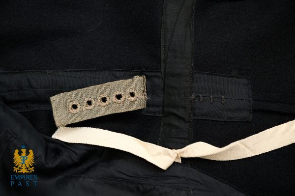 Close-up of a black coat interior showing a beige strap with five punched holes and a beige belt, Empires Past logo bottom-left.