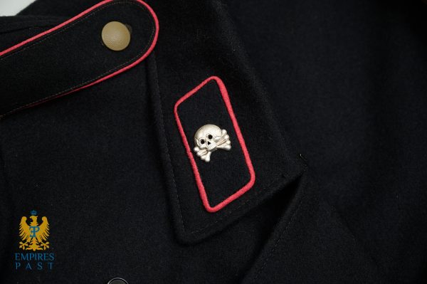 Close-up of a black military-style coat shoulder with red piping and a shiny silver skull-and-crossbones badge on a rectangular patch with rounded corners.