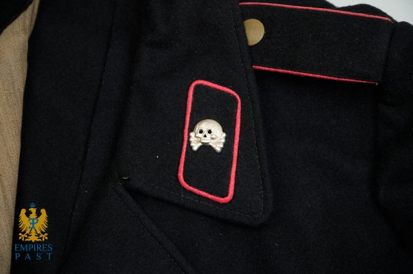 Close-up of a dark military jacket with red piping and a skull-and-crossbones pin on the chest pocket.