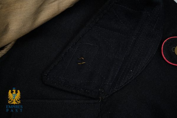 Close-up of a dark navy uniform jacket with visible stitching, a red-edged circular patch on the chest, and beige fabric at the top.