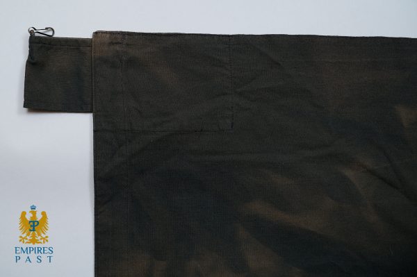 Close-up of dark fabric with a small rectangular stitched pocket near the top left and a metal loop at the edge; Empires Past logo appears in the bottom-left corner.