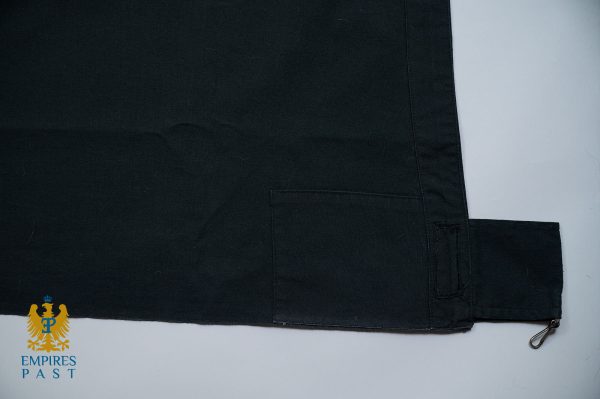Close-up of dark fabric with a rectangular stitched pocket and a small fabric tab on the lower edge; a metal zipper pull is visible at the right edge.