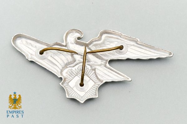 Wing-shaped metal badge with two holes threaded by a brown cord, lying on a pale gray surface; Empires Past logo visible in the lower-left corner.