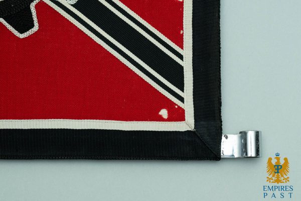 Close-up of a red flag corner with a black border and white trim, attached to a metal clip on a gray background, with the Empire Past logo in the bottom-right.