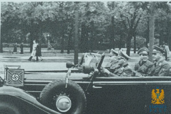 Open-top military car with two uniformed officers waving during a Nazi-era parade; swastika flag visible on the vehicle's front.