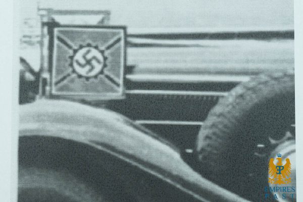 Square sign with a swastika emblem mounted on a surface, in a black-and-white photo; surrounding details are blurred, suggesting a historical Nazi insignia context.