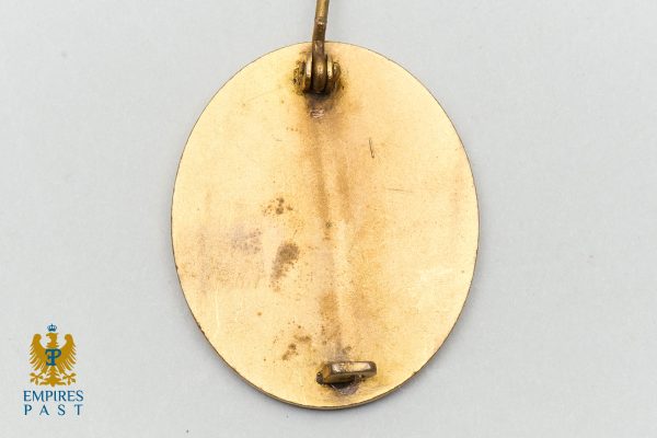 Empires Past 202517294 - Empires Past Gold oval pendant back with a small loop at the top, showing patina marks on a light blue surface; Empires Past logo visible in the lower-left corner.