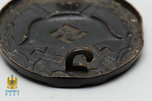 Empires Past 202517362 - Empires Past Close-up of a dark circular metal badge or seal with a curved hook in the center and a worn, textured surface; Empries Past logo in corner.