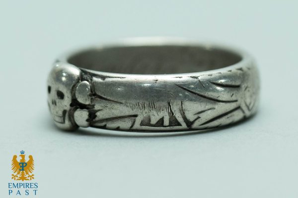 Close-up of a silver ring with a skull motif and carved etchings along the band on a pale surface