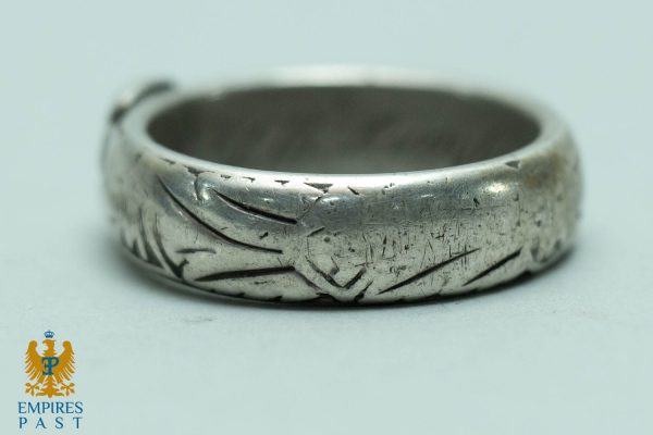 Close-up of a worn silver ring with a leaf-like engraved pattern and many scratches on a pale background.