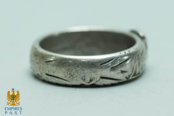 Close-up of a worn silver ring with diagonal engraved leaf-like patterns and surface scratches on a light background.