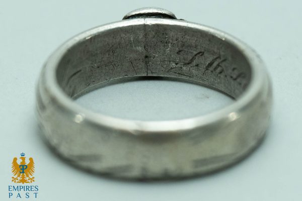 Close-up of a silver ring with a scratched inner surface and faint engraving, logo in bottom-left corner.
