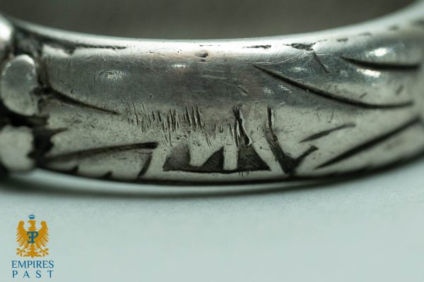 Macro shot of a worn silver ring showing deep scratches and engraved marks; Empires Past logo visible in the lower-left corner.
