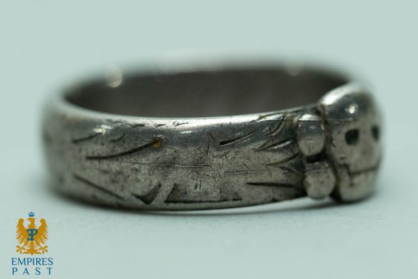 Close-up of a scratched silver ring with engraved linear patterns, featuring a small hinged area, placed on a pale surface with the Empires Past logo in the corner.
