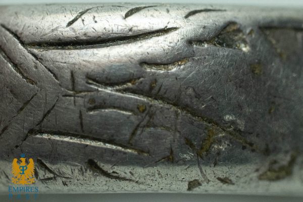 Close-up of a scratched metal surface with deep gouges; bottom-left shows a small blue-and-gold Empires Past crest.