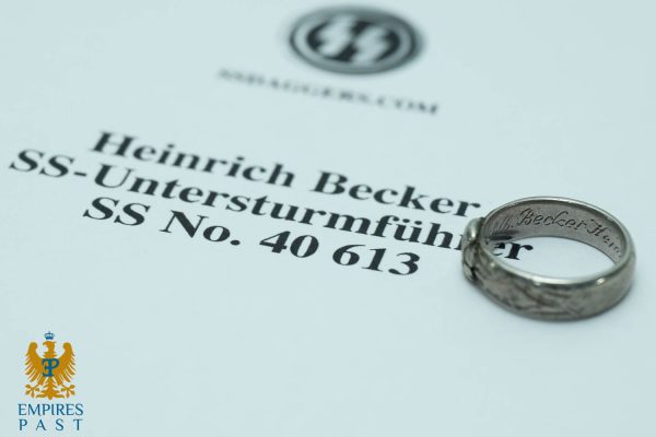 Close-up of a historical document page with the name Heinrich Becker and SS No. 40613, a silver ring resting on the text; Empires Past logo bottom-left.